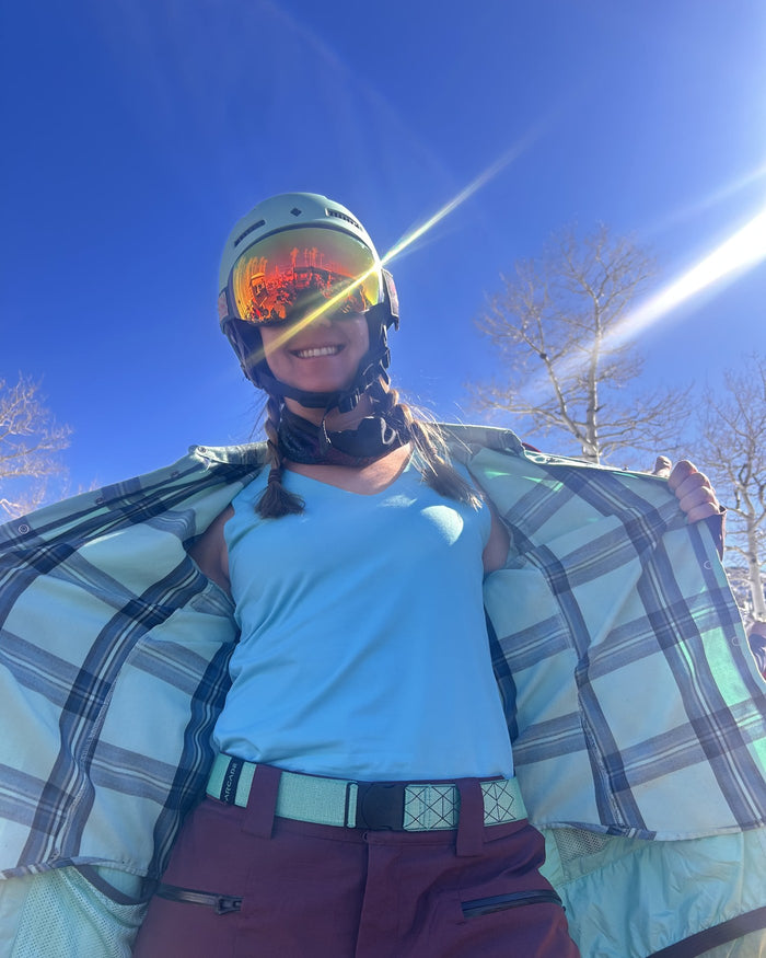 Woman on the mountain in a ski helmet and goggles, holding open a plaid flannel over a light blue JudyP sleeveless top